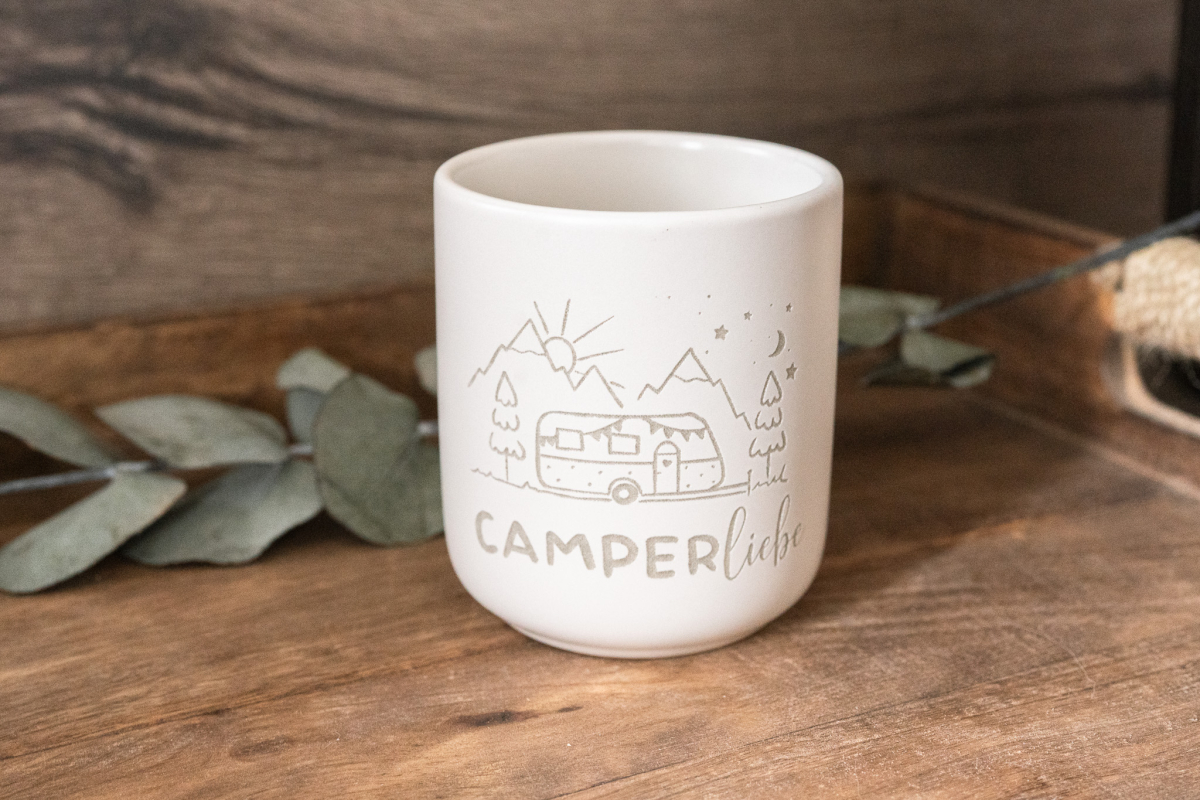 Matte weiße Keramiktasse mit Gravur „Camperliebe“ und Camping-Motiv (Wohnwagen, Berge) auf Holzuntergrund, Eukalyptus im Hintergrund.
